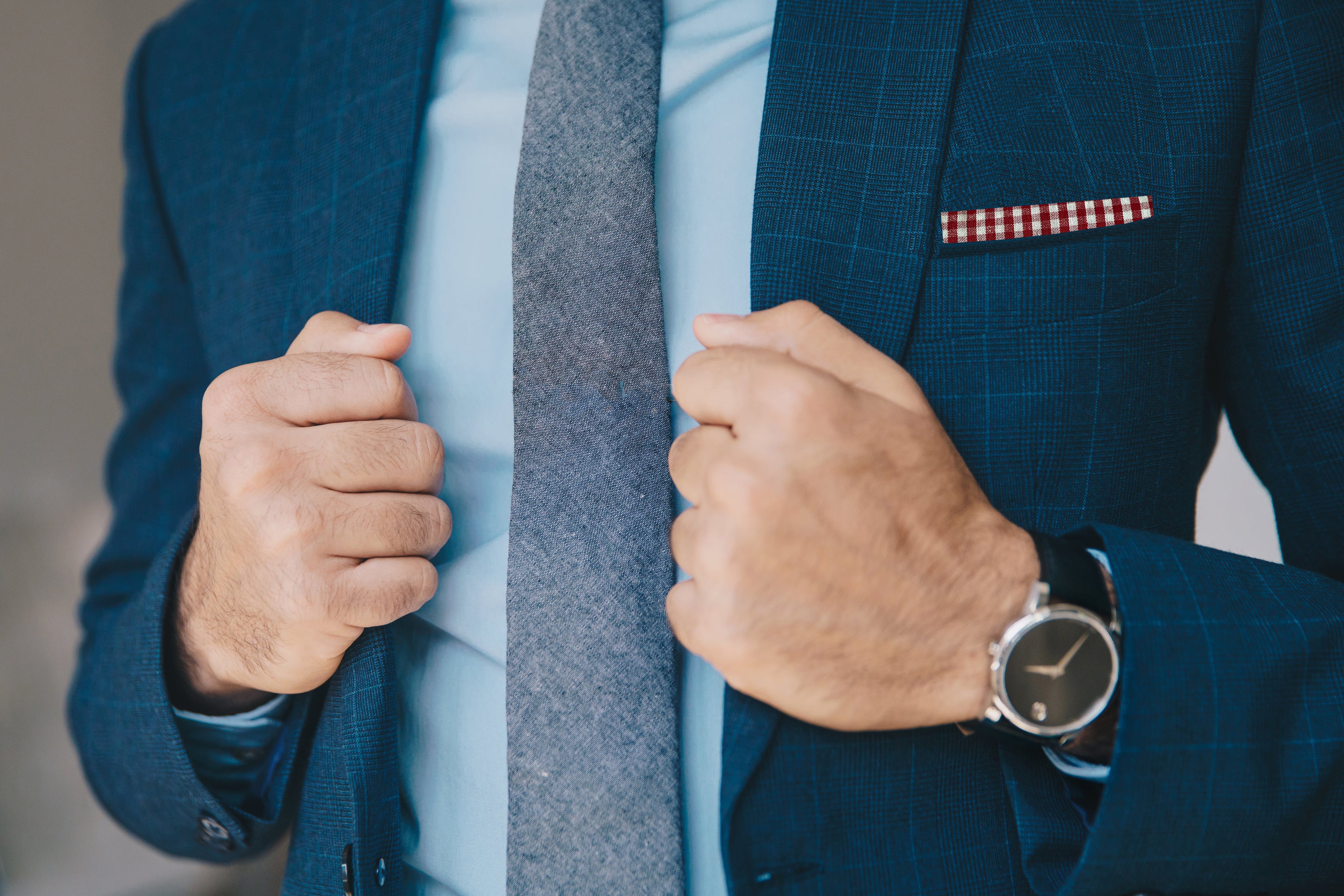 Person with a gray tie and red pocket square with a blue suit jacket and watch.